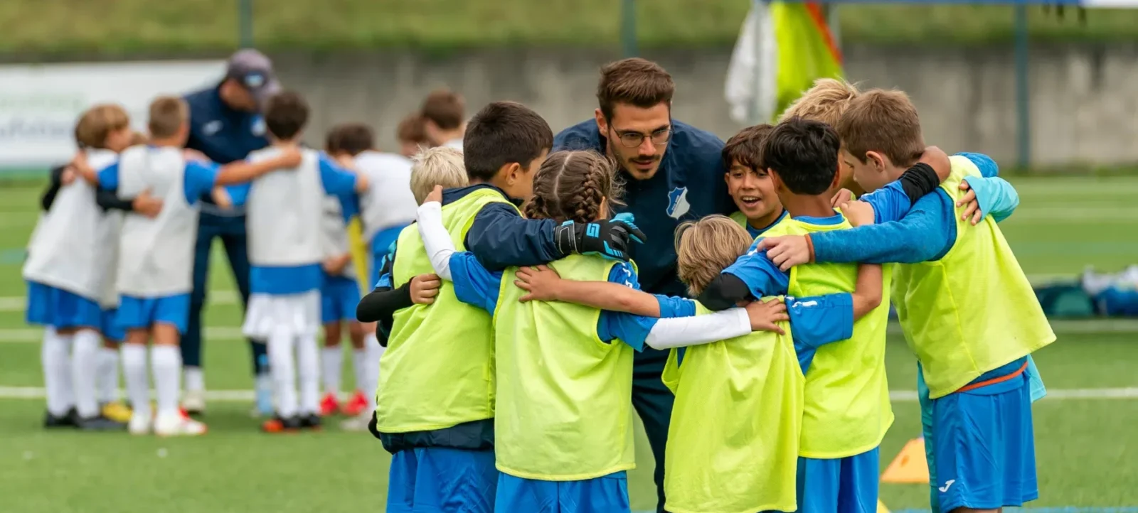 Eine Gruppe von Kindern und ein Trainer in blauen und gelben Trikots umarmen sich auf einem Fußballfeld. Die Kinder scheinen sich zu freuen und sind in einer engen Gruppe zusammengeschlossen. Im Hintergrund sind weitere Kinder und ein Trainer zu sehen, die ebenfalls in einer ähnlichen Gruppe stehen. Die Szene findet auf einem grünen Rasenplatz statt, und die Atmosphäre wirkt fröhlich und gesellig.