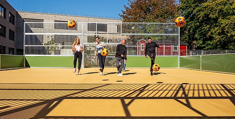Vier Personen spielen auf einem Bolzplatz Fußball. Sie sind in Bewegung und kicken gleichzeitig vier gelb-schwarze Bälle. Im Hintergrund ist ein Gebäude mit großen Fenstern und ein Zaun zu sehen. Die Szene spielt bei Tageslicht unter einem klaren Himmel.
