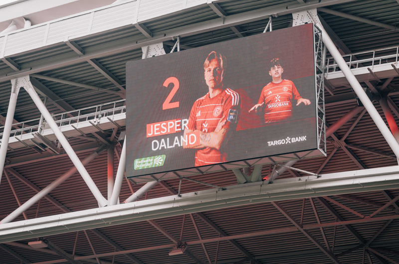 Große Stadionanzeigetafel mit zwei Fußballspielern im Trikot des 1. FC Köln. Die Tafel zeigt die Nummer 2 und den Namen "Jesper Daland". Die Spieler machen eine Gebärdensprache-Geste. Im Hintergrund ist das Dach des Stadions zu sehen.