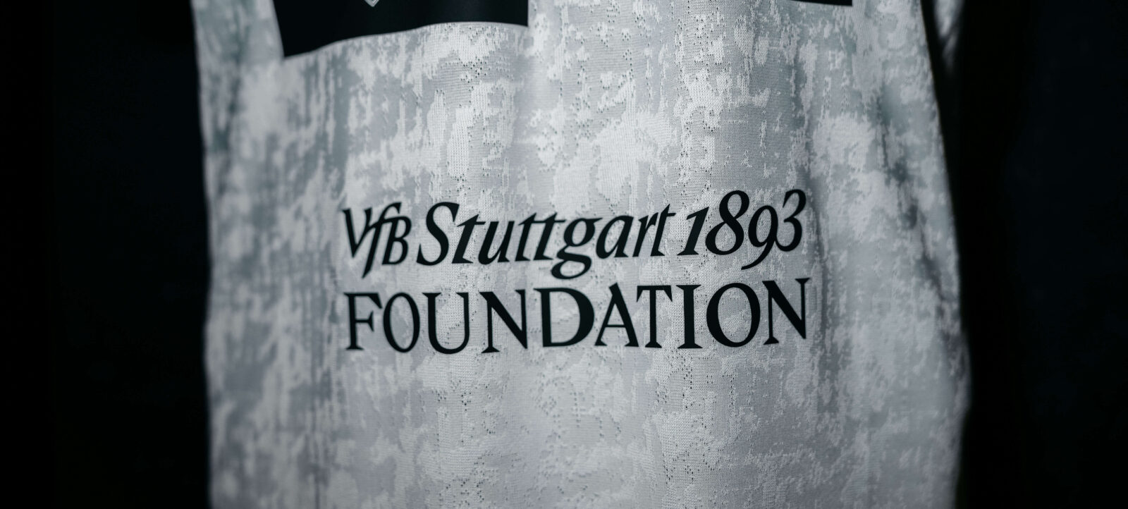 Ein graues Trikot mit dem Aufdruck "VfB Stuttgart 1893 Foundation" und zwei schwarzen Schulterstücken, die das Vereinslogo tragen.
