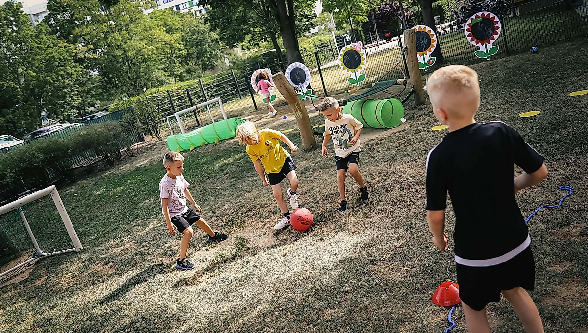 Kinder spielen auf einem Sportplatz im Freien. Sie sind in verschiedenen Aktivitäten wie Fußballspielen und Hindernisläufen beschäftigt. Der Platz ist mit grünen Hindernissen und bunten Blumen-Deko-Elementen ausgestattet. Die Kinder tragen Sportkleidung und sind in einer natürlichen, umzäunten Umgebung mit Bäumen und Gras.