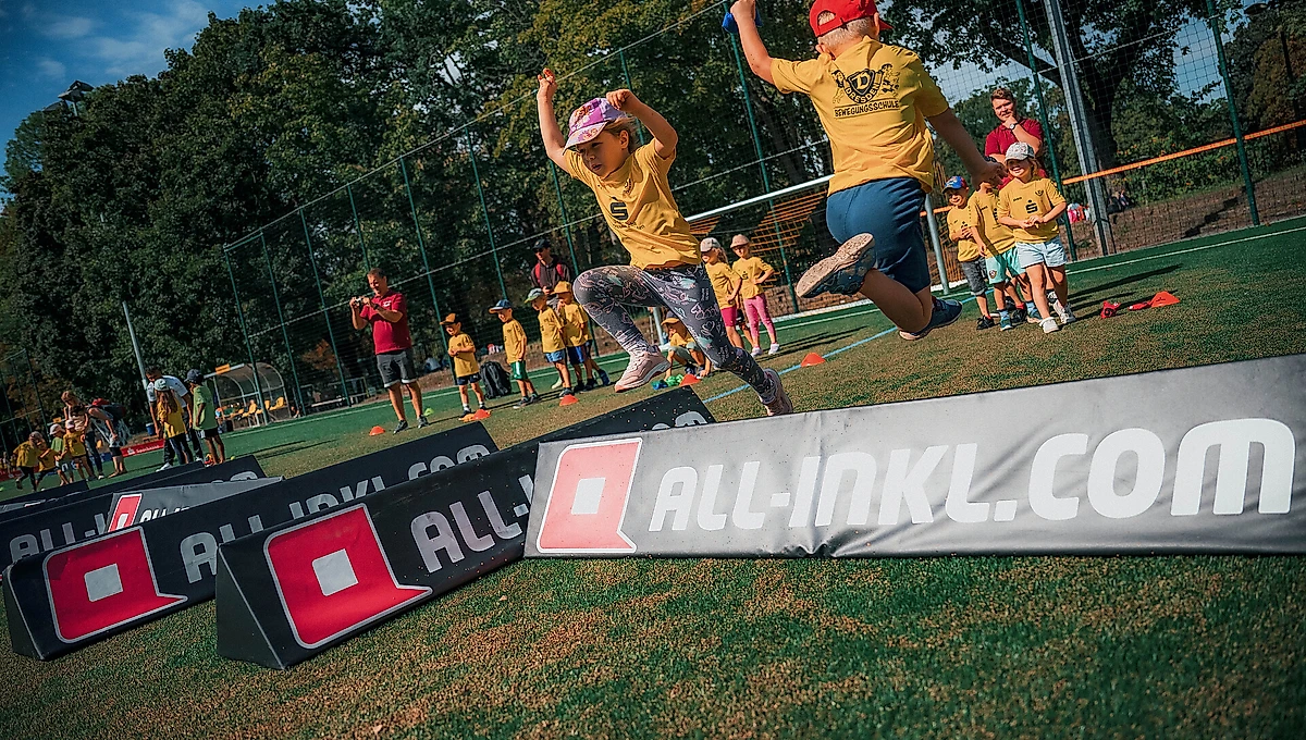 Kinder und Betreuer in gelben T-Shirts nehmen an einem Bewegungstag teil. Sie springen über Hindernisse auf einem Fußballfeld, im Hintergrund sind weitere Teilnehmer und Bäume zu sehen. Im Vordergrund ist ein Werbebanner mit dem Logo "all-inkl.com" zu erkennen.