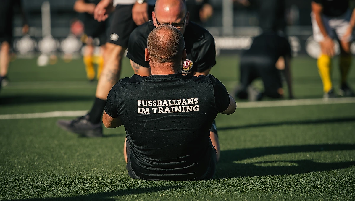 Ein Mann sitzt auf dem Rasen eines Fußballfeldes und beobachtet ein Training. Er trägt ein schwarzes T-Shirt mit der Aufschrift "FUSSBALLFANS IM TRAINING" auf dem Rücken. Im Hintergrund sind Fußballspieler in gelben Hosen und schwarze Schiedsrichter zu sehen.