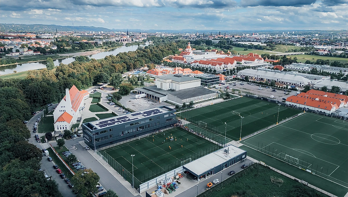 Luftaufnahme eines Fußballtrainingsgeländes mit mehreren Rasenplätzen und einem modernen Gebäude. Im Hintergrund ist eine Stadt mit einem Fluss und historischen Gebäuden zu sehen.
