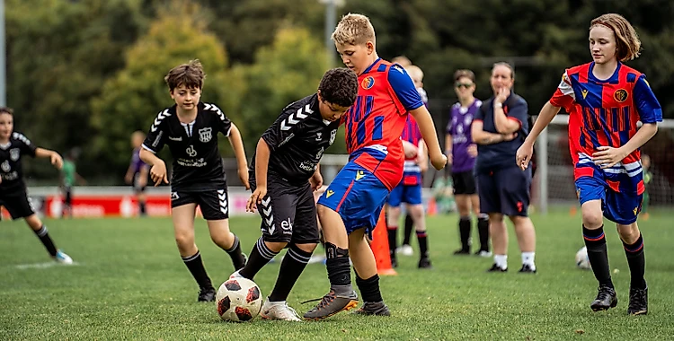 Jugendliche Fußballer in einem Inklusionsspiel. Zwei Spieler in schwarzen Trikots und zwei Spieler in roten und blauen Trikots umkämpfen sich den Ball auf einem Rasenplatz. Im Hintergrund sind weitere Spieler und Betreuer zu sehen.