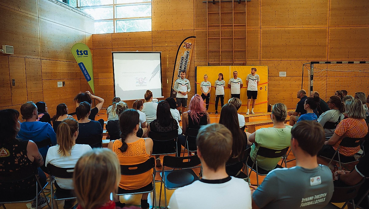 Eine Gruppe von Menschen sitzt in einem Raum und hört einer Präsentation zu. Auf der Bühne stehen vier Personen, die vor einem Projektor stehen, auf dem ein Bild zu sehen ist. Die Personen auf der Bühne tragen T-Shirts mit dem Logo der Fußballschule. Im Raum sind zwei Banner mit dem Logo der Fußballschule und einem anderen Logo zu sehen. Der Raum hat holzverkleidete Wände und große Fenster, die natürliches Licht hereinlassen.