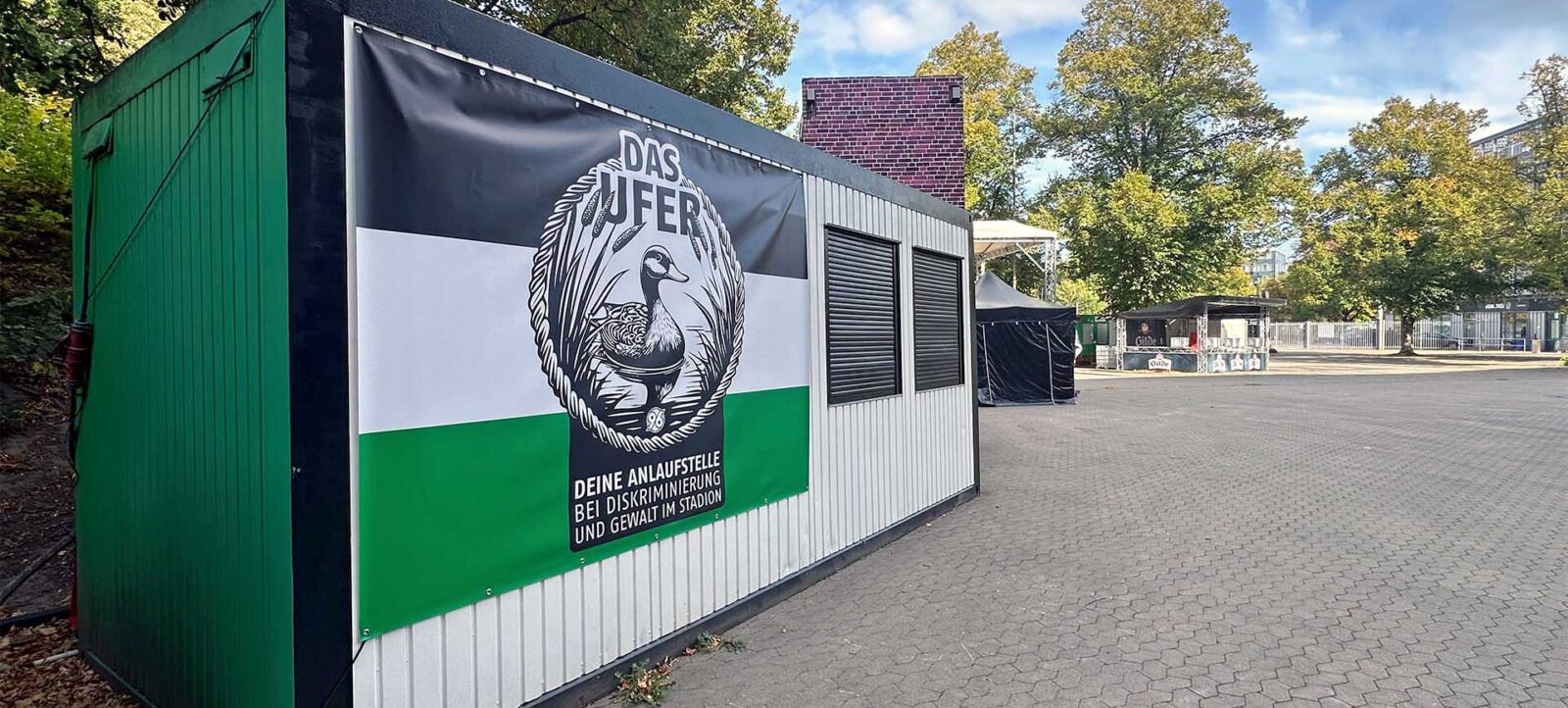 Ein grün-weißer Container mit einem Schild, das ein Vogelmotiv und den Text "DEINE ANLAUFSTELLE BEI DISKRIMINIERUNG UND GEWALT IM STADION" zeigt. Der Container steht auf einem gepflasterten Platz mit Bäumen und weiteren Zelten im Hintergrund.