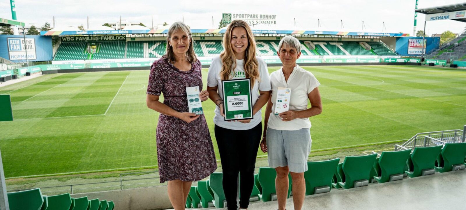 Drei Frauen stehen auf den Tribünen eines Fußballstadions. Die Frau in der Mitte hält einen Scheck über 2.000 Euro in den Händen, während die beiden Frauen links und rechts jeweils ein Informationsblatt halten. Im Hintergrund ist das Spielfeld und die Tribüne des Stadions zu sehen.