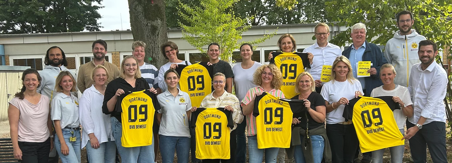 Eine Gruppe von Menschen steht vor einem Gebäude und hält gelbe Trikots mit der Aufschrift "Dortmund 09 BVB bewegt" in die Kamera. Die Personen lächeln und wirken fröhlich. Im Hintergrund sind Bäume und ein Teil des Gebäudes zu sehen.