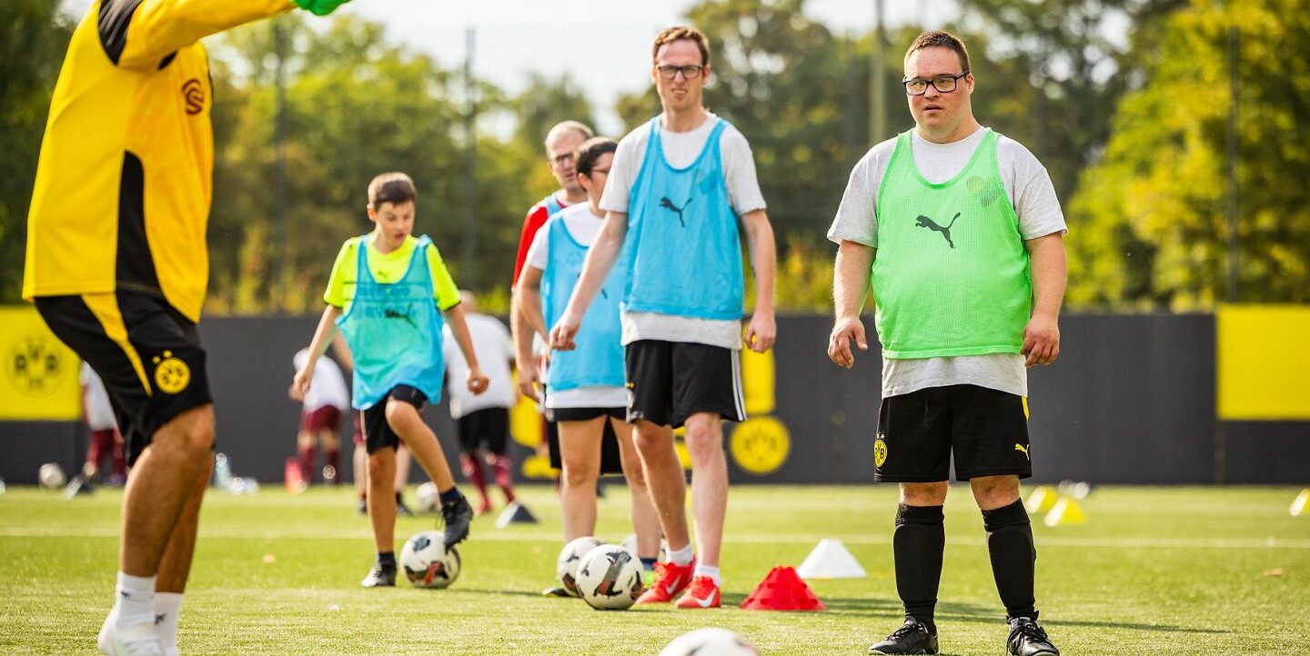 Ein Fußballtrainer in einem gelben Trikot mit schwarzem Design gibt Anweisungen an eine Gruppe von Teilnehmern, die bunte Trainingswesten tragen. Die Gruppe steht auf einem Fußballfeld und übt mit Bällen und Hütchen. Die Teilnehmer scheinen sich auf ein Training oder eine Übungseinheit zu konzentrieren.
