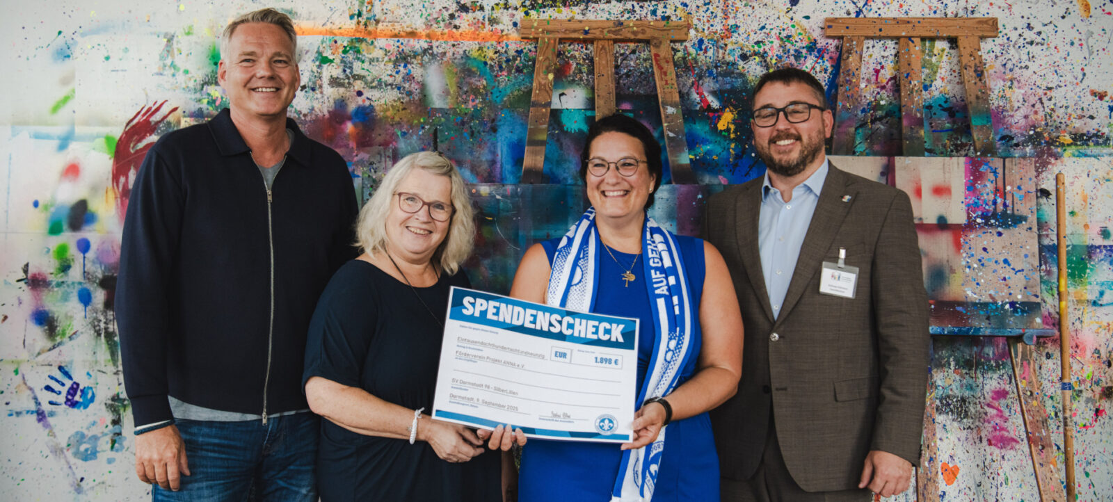Vier Personen stehen vor einem farbenfrohen Hintergrund und halten gemeinsam einen großen Scheck mit der Aufschrift "SPENDENSCHECK" und einem Betrag von 1.098 Euro. Die Person in der Mitte trägt eine blaue Schärpe und hält den Scheck, während die anderen drei Personen lächeln und sich über die erfolgreiche Spendenübergabe freuen.