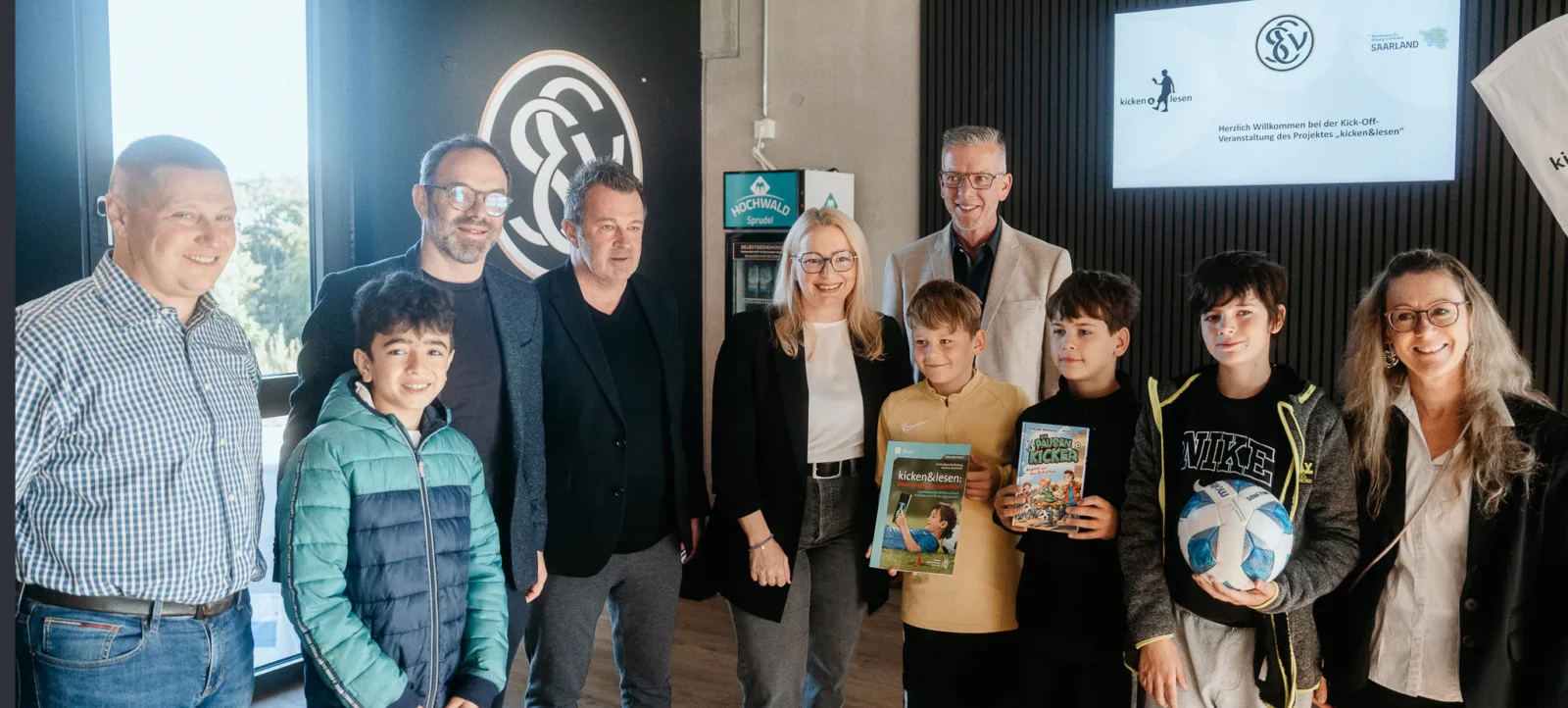 Eine Gruppe von Erwachsenen und Kindern steht vor einem Hintergrund mit dem Logo des 1. FC Saarbrücken und einem Schild, das das Projekt "kicken & lesen" ankündigt. Die Kinder halten Bücher und einen Fußball in den Händen. Alle lächeln und scheinen sich über den Kick-Off des Projekts zu freuen.