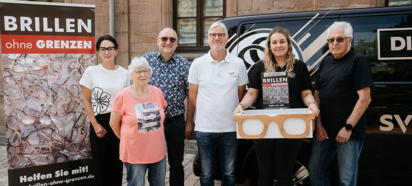 Eine Gruppe von fünf Personen steht vor einem Transporter und einem Banner mit der Aufschrift "Brillen ohne Grenzen". Eine der Personen hält eine große Box mit der Aufschrift "Brillen ohne Grenzen" und einem Bild von Brillen. Das Banner zeigt viele Brillen und den Aufruf "Helfen Sie mit!". Die Personen lächeln und scheinen sich für eine wohltätige Aktion zu engagieren.
