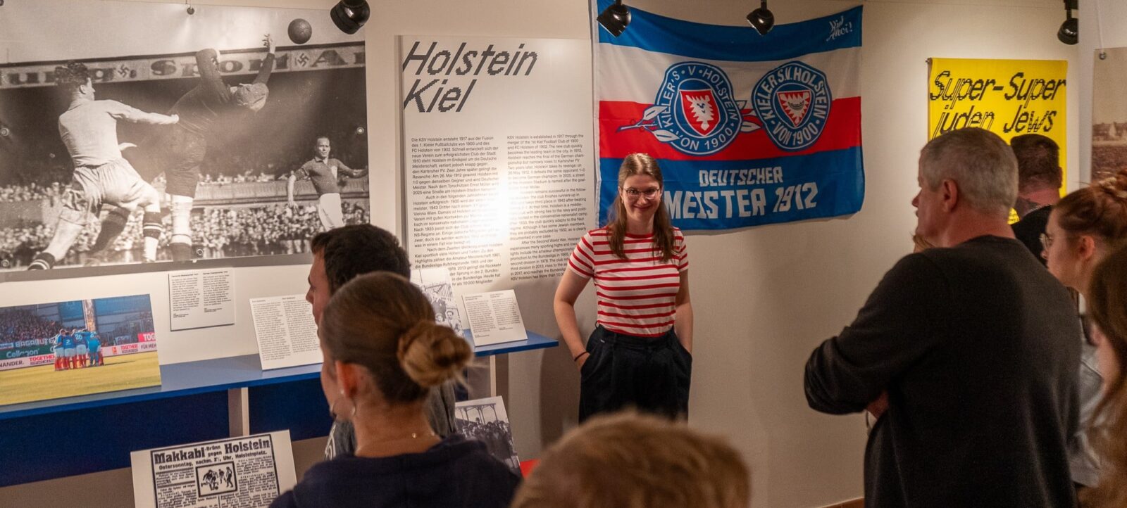 Eine Gruppe von Menschen steht vor einer Ausstellung über den Fußballverein Holstein Kiel. Eine Frau in einem rot-weiß gestreiften T-Shirt steht vor einer Informationswand und erklärt die Geschichte des Vereins. Die Wand zeigt historische Fotos, eine blau-weiße Fahne mit dem Vereinswappen und den Aufschrift "Deutscher Meister 1912" sowie ein gelbes Plakat mit der Aufschrift "Super-Super Juden Jutta". Die Besucher scheinen aufmerksam zuzuhören und die Ausstellung zu betrachten.