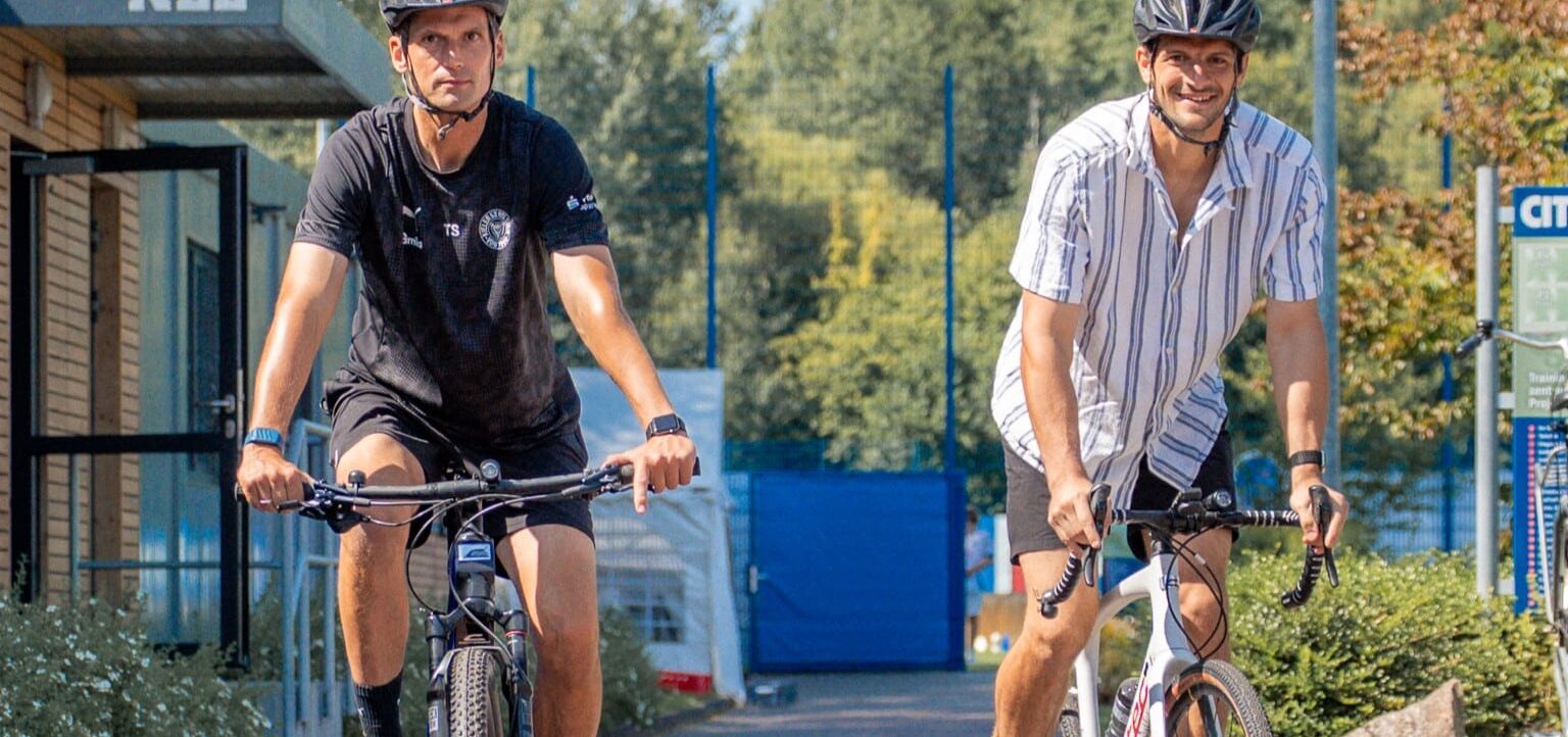 Zwei Männer fahren Fahrrad auf einem Weg neben einem Gebäude mit der Aufschrift "NLZ". Beide tragen Helme und Sportkleidung. Der Hintergrund zeigt grüne Bäume und Büsche.