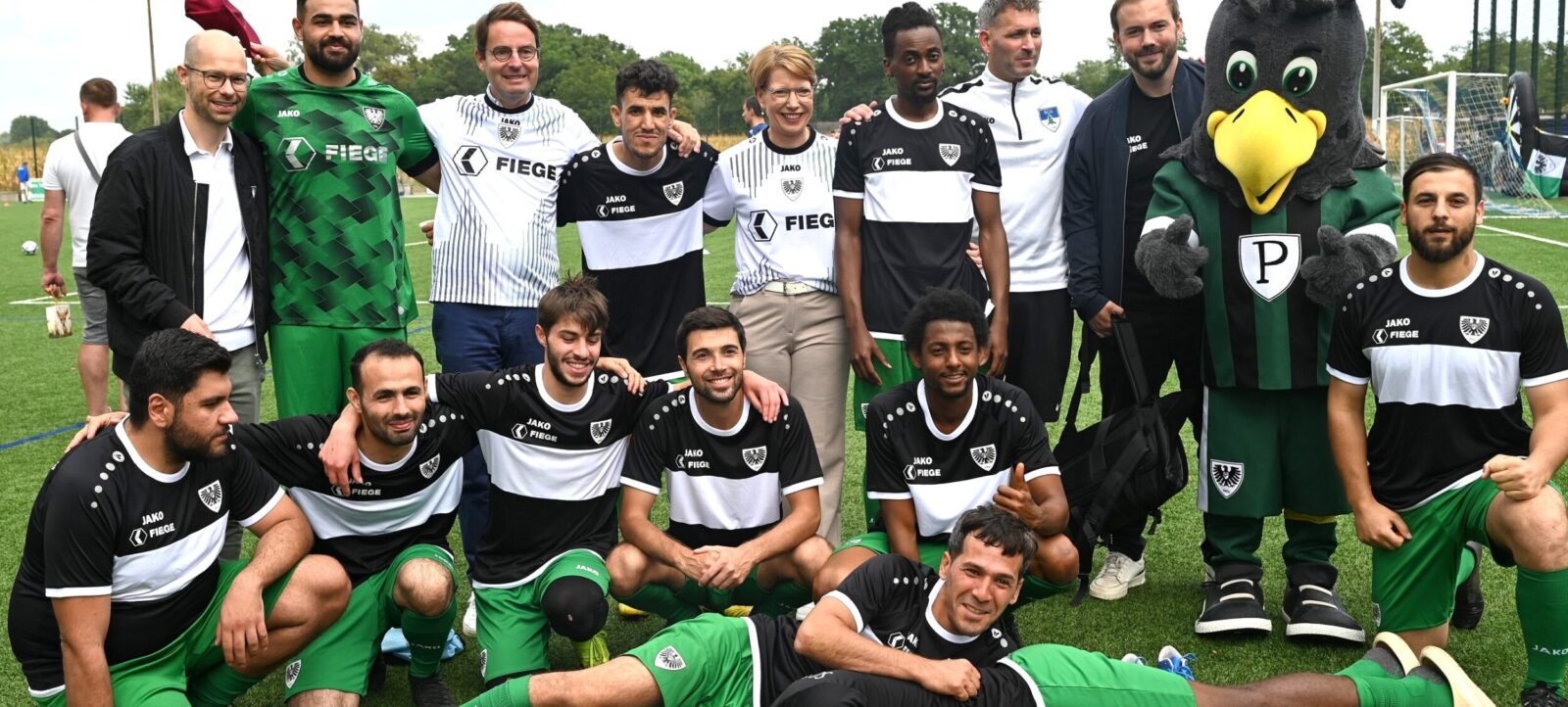 Eine Gruppe von Fußballspielern und Betreuern posiert für ein Teamfoto auf einem Rasenplatz. Die Spieler tragen schwarze und grüne Trikots mit weißen Streifen und dem Logo des Vereins. Ein Maskottchen in Form eines Vogels mit grünen und schwarzen Federn und einem weißen "P" auf der Brust steht mit im Bild. Die Spieler sind in verschiedenen Posen, einige sitzen, andere knien oder liegen auf dem Boden, und alle lächeln in die Kamera. Im Hintergrund sind Fußballtore und eine grüne Landschaft zu sehen.