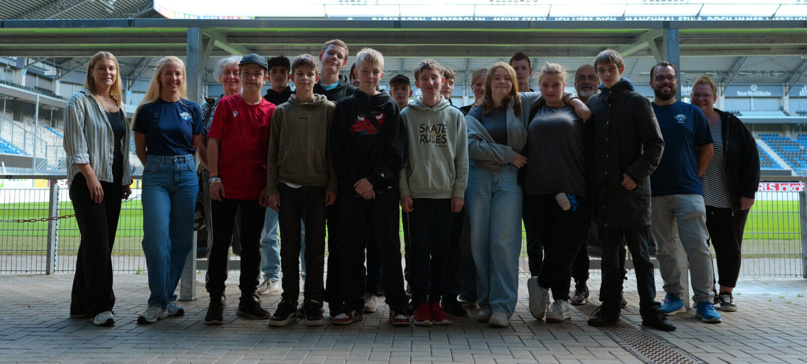 Eine Gruppe von Menschen steht vor einem Fußballstadion und lächelt in die Kamera. Die Gruppe besteht aus einer Mischung von Erwachsenen und Jugendlichen, die unterschiedlich gekleidet sind. Im Hintergrund sind die Sitzplätze des Stadions und ein Teil des Spielfelds zu sehen.