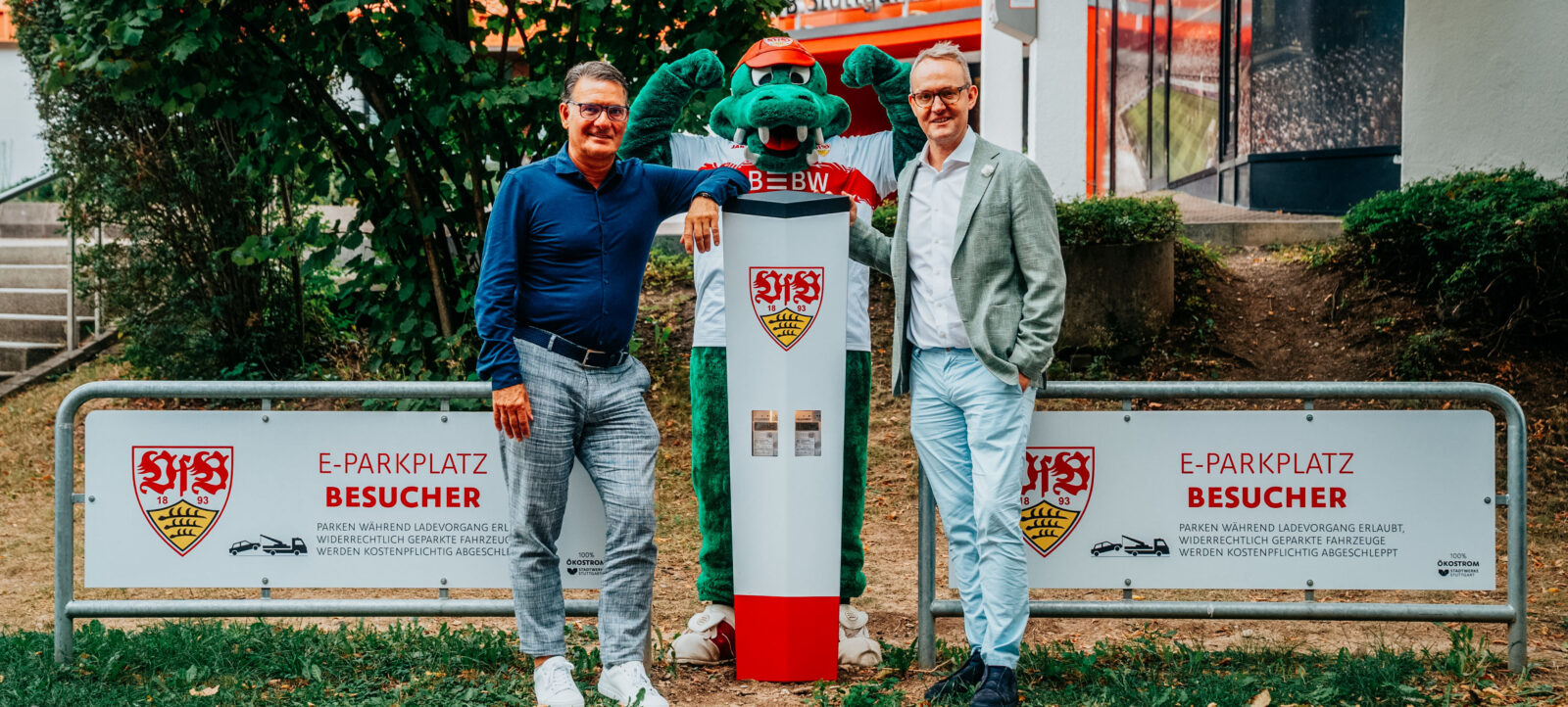 Zwei Männer und eine Maske des Vereinsmaskottchens stehen vor einer E-Ladesäule des Vereins VfB Stuttgart. Die Ladesäule ist mit dem Vereinswappen und dem Text "E-PARKPLATZ BESUCHER" beschriftet.