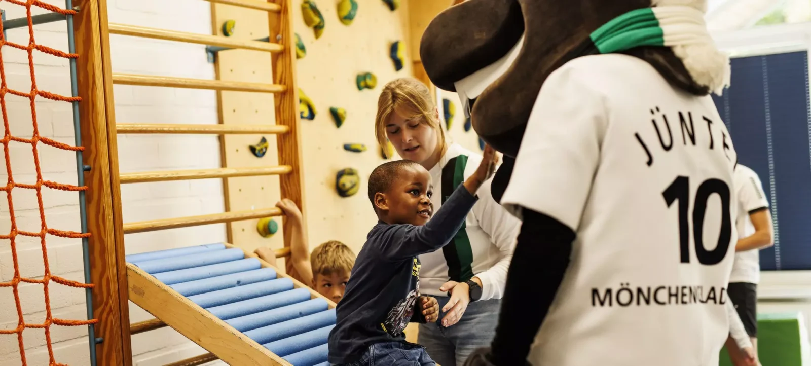 Kinder und eine Betreuerin in einem Kletterraum. Die Kinder klettern an einer Kletterwand mit Sprossen und einem Netz. Ein Kind trägt ein T-Shirt mit der Aufschrift "Jünta 10" und "Mönchenlad". Die Betreuerin unterstützt die Kinder beim Klettern.