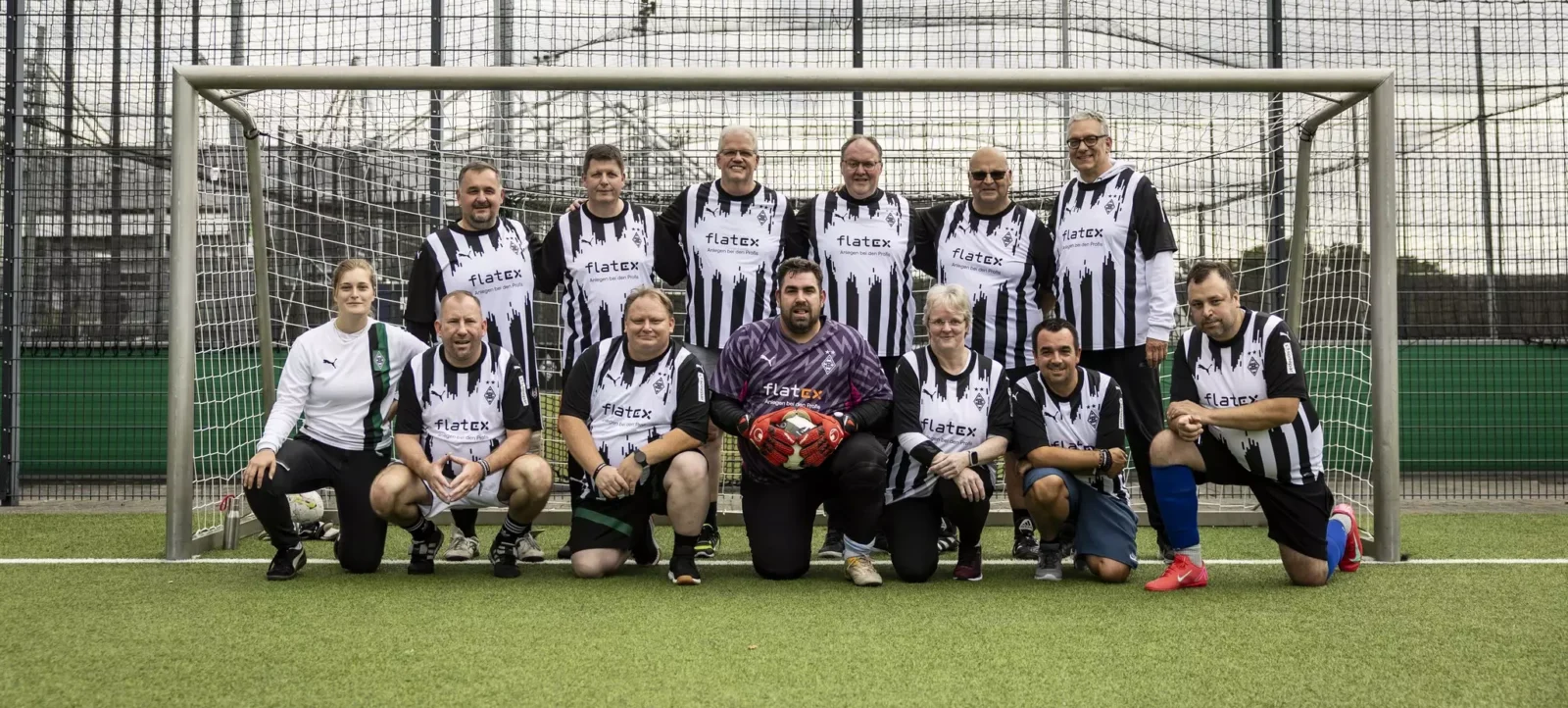 Eine Gruppe von Menschen in Fußballtrikots posiert vor einem Tor auf einem Fußballfeld. Die Trikots sind schwarz-weiß gestreift mit dem Logo von Flatex und einem abstrakten Design. Die Gruppe besteht aus Männern und Frauen, die sowohl stehen als auch knien. Einige tragen Schienbeinschoner und Fußballschuhe, und eine Person hält einen Torwarthandschuh. Im Hintergrund ist ein Netz und ein Zaun zu sehen.