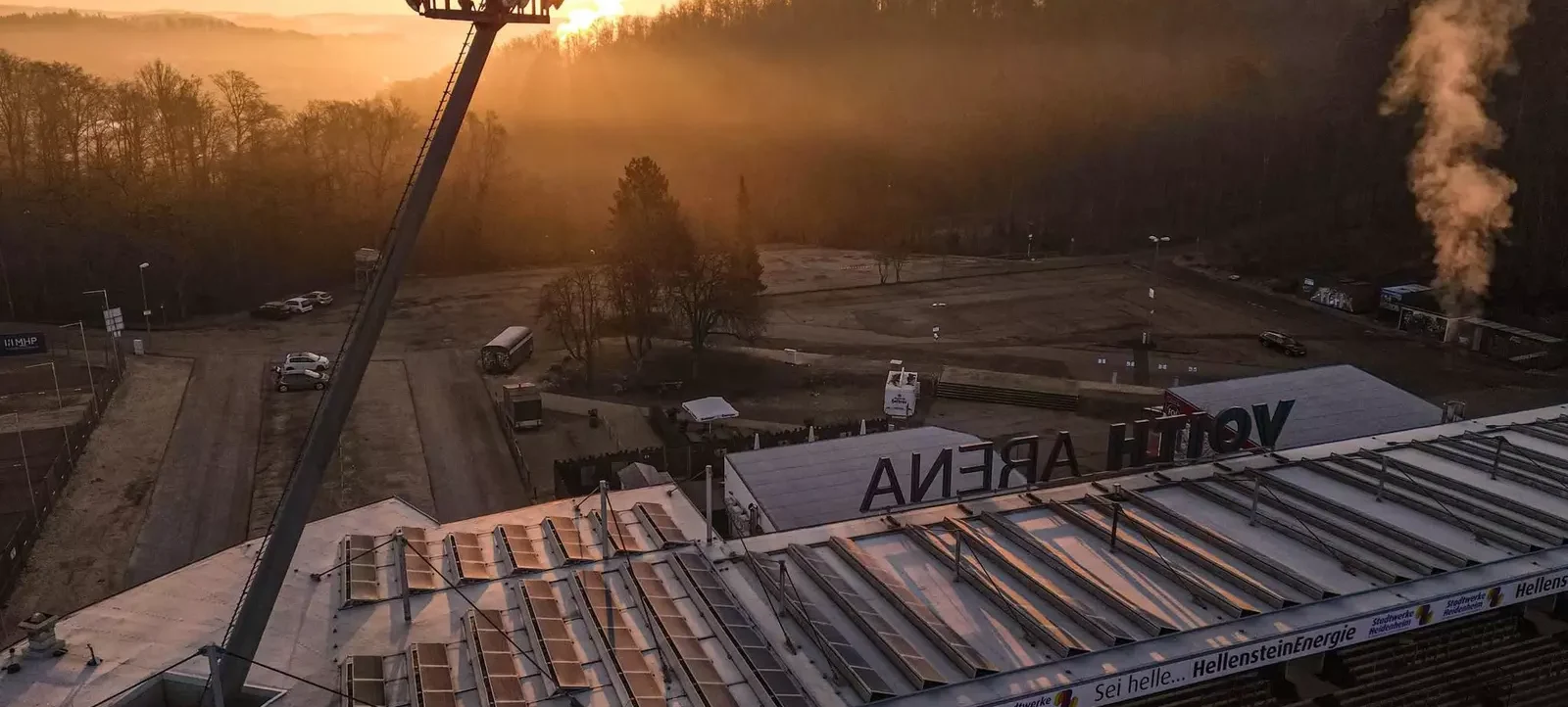 Ein Stadion bei Sonnenaufgang mit leichten Nebelschwaden. Die Sonne geht hinter einem Flutlichtmast auf, der sich über das Dach des Stadions erstreckt. Auf dem Dach des Stadions ist das Wort 'ARENA' zu sehen. Im Hintergrund sind Bäume und ein Parkplatz mit einigen Fahrzeugen zu erkennen.