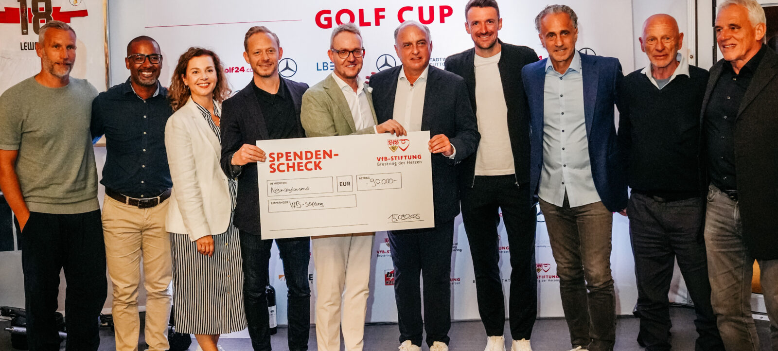 Eine Gruppe von Menschen hält einen großen Spendenscheck im Wert von 30.000 Euro für die VfB-Stiftung bei einem Golf Cup Event. Die Teilnehmer lächeln und stehen vor einem Banner mit dem Logo des VfB Stuttgart und dem Text "GOLF CUP".
