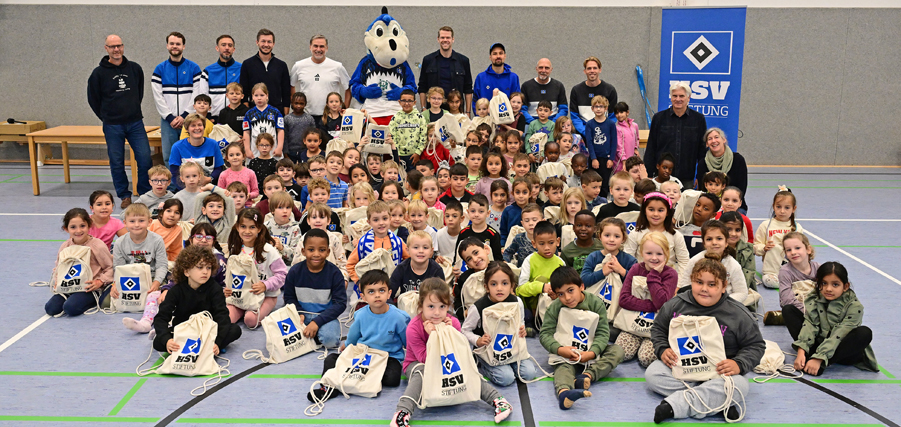 Eine große Gruppe von Kindern und Erwachsenen in einer Sporthalle, darunter auch das HSV-Maskottchen. Die Kinder sitzen auf dem Boden und halten weiße Stoffbeutel mit dem HSV-Logo. Die Erwachsenen stehen im Hintergrund und lächeln in die Kamera. Die Szene findet im Rahmen der Übergabe von Schulstarterpaketen an der Fridtjof-Nansen-Schule statt.