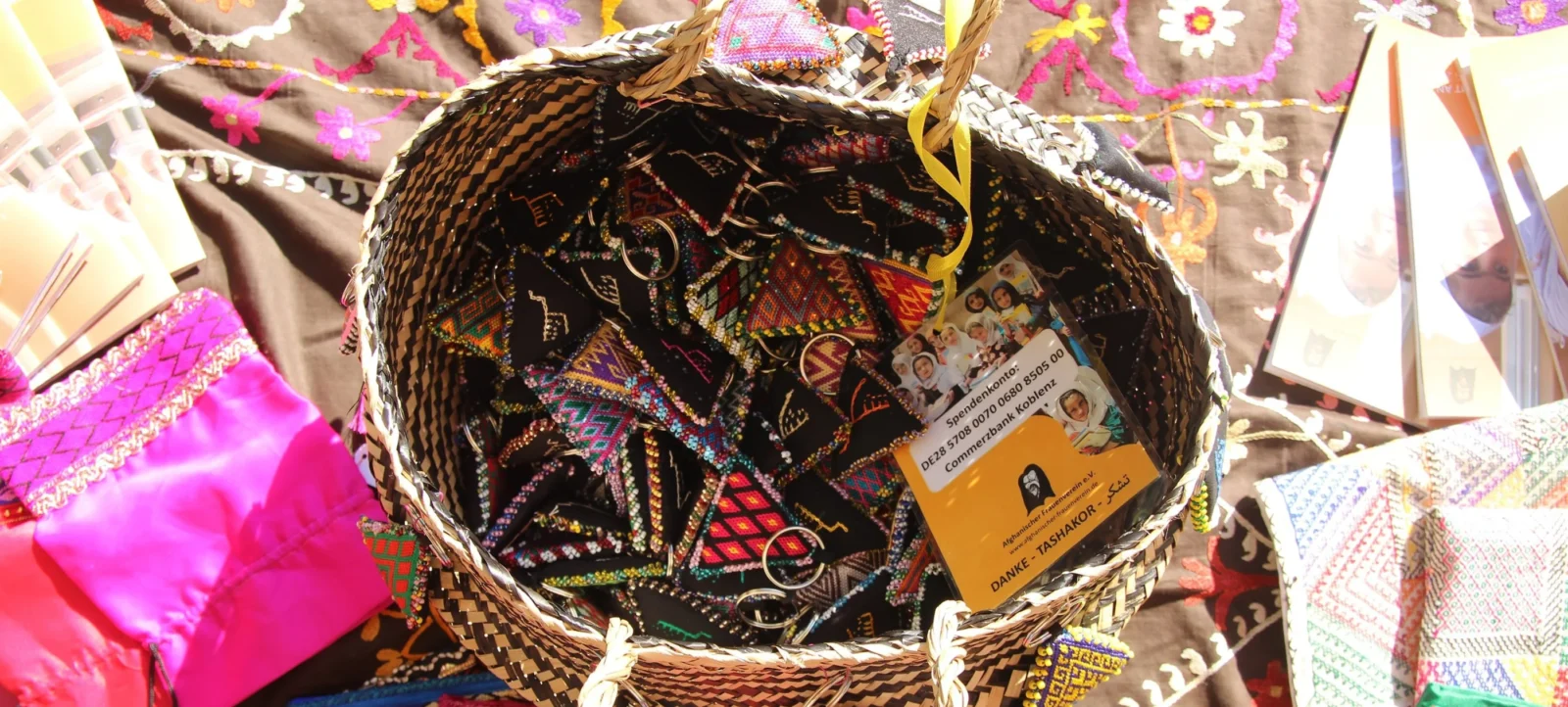 Ein geflochtener Korb gefüllt mit farbenfrohen, handgefertigten Textilien und einem Dankesschild. Der Korb ist mit bunten Stoffen und Stickereien verziert und enthält auch ein Foto mit mehreren Personen sowie einen Spendenhinweis.