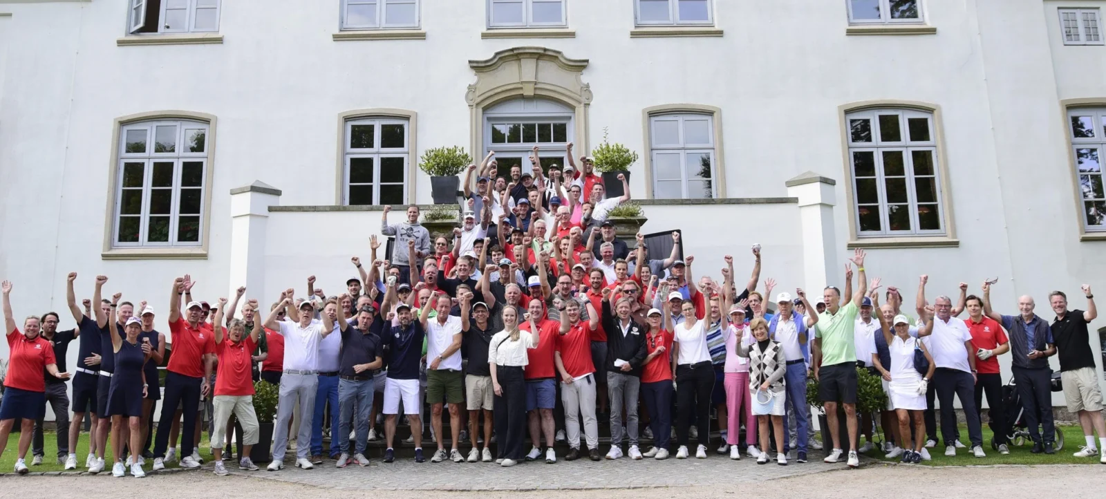 Eine große Gruppe von Menschen steht vor einem weißen Gebäude mit mehreren Fenstern und einem zentralen Eingang. Die Menschen sind in mehreren Reihen angeordnet und heben ihre Arme in die Luft, was auf eine feierliche oder motivierte Stimmung hindeutet. Die Gruppe besteht aus Männern und Frauen, die unterschiedliche Kleidung tragen, darunter Polo-Shirts, T-Shirts und Sportkleidung. Das Gebäude im Hintergrund hat einen klassischen architektonischen Stil mit großen Fenstern und einem zentralen Eingang, der von zwei Säulen flankiert wird. Die Szene wirkt wie ein Gruppenfoto oder eine Teamaufstellung, möglicherweise im Rahmen eines sportlichen oder gesellschaftlichen Ereignisses.