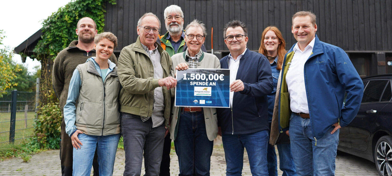 Eine Gruppe von sieben Personen steht vor einem Holzgebäude und lächelt in die Kamera. Zwei der Personen in der Mitte halten gemeinsam ein Schild, auf dem "1.500,00€ SPENDE AN" steht. Die Gruppe scheint sich über eine erfolgreiche Spendenaktion zu freuen.