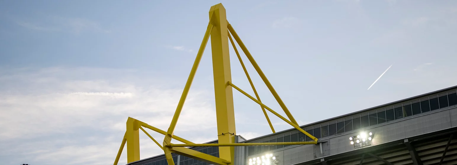 Gelbe Stahlträger eines Fußballstadions vor einem klaren Himmel. Im Hintergrund ist ein Teil der Stadionstruktur mit Fenstern und Flutlichtanlagen zu sehen.