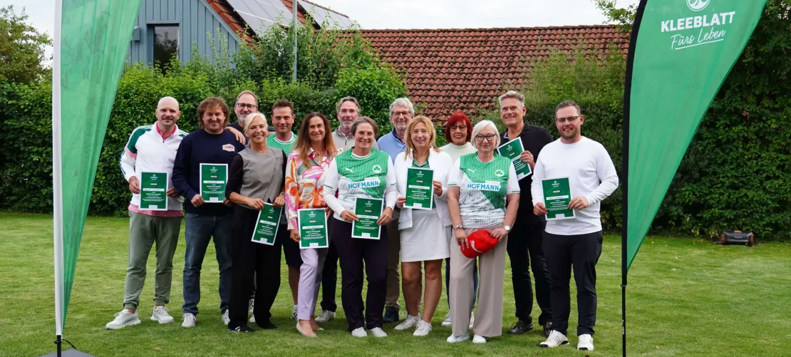 Eine Gruppe von Menschen steht auf einem Rasen vor einem Gebäude mit Solaranlage auf dem Dach. Sie halten grüne Urkunden in den Händen und lächeln in die Kamera. Im Hintergrund sind grüne Banner mit dem Logo "Kleeblatt Fürs Leben" zu sehen.