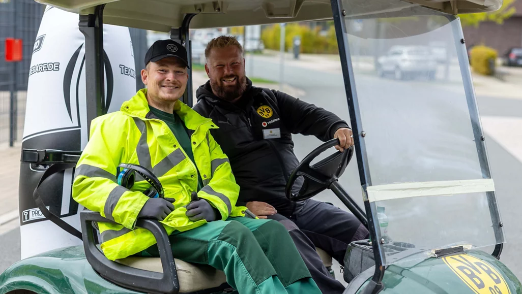 Zwei Mitarbeiter in Arbeitskleidung sitzen in einem Golfwagen. Der eine trägt eine gelbe Sicherheitsjacke und eine schwarze Kappe, der andere eine schwarze Jacke mit einem Vereinslogo und einen Bart. Sie lächeln und scheinen sich zu unterhalten. Der Golfwagen steht auf einem Parkplatz oder einer Straße.