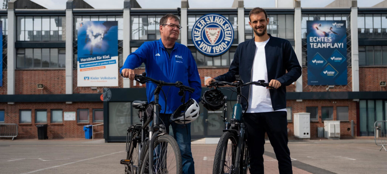 Zwei Männer stehen vor dem Stadion des Kieler SV Holstein von 1900 und halten Fahrräder. Der Mann links trägt eine blaue Jacke mit der Aufschrift "Kiel" und "Scandinavian City", während der Mann rechts ein weißes T-Shirt und eine dunkle Jacke trägt. Im Hintergrund sind Plakate mit Werbung für die Kieler Volksbank und das Motto "Echtes Teamplay!" zu sehen.