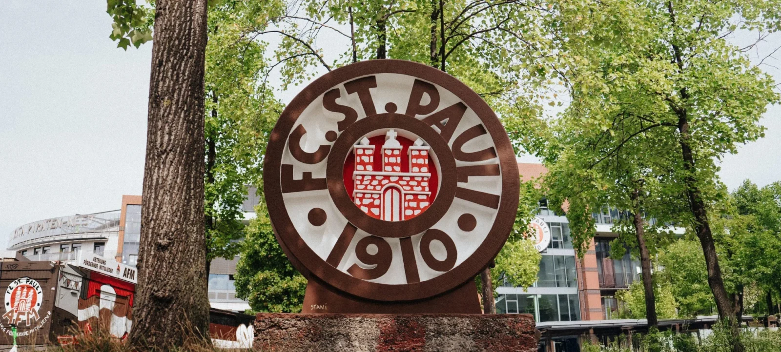 Ein großes, rundes Steinwappen des FC St. Pauli mit dem Vereinslogo und der Jahreszahl 1910, umgeben von Bäumen und modernen Gebäuden im Hintergrund.