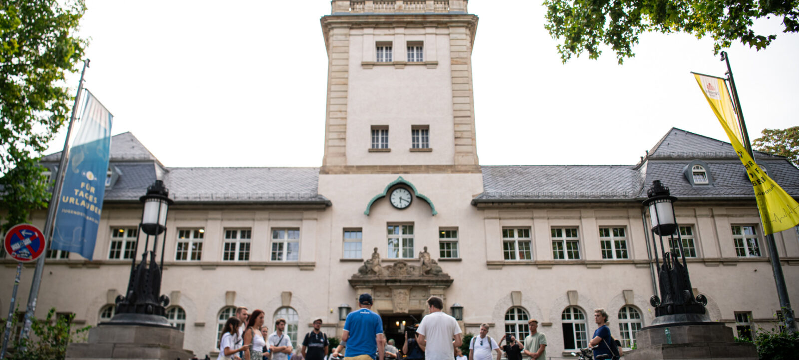 Ein historisches Gebäude mit einem Uhrturm, vor dem sich eine Gruppe von Menschen versammelt hat. Einige Personen stehen in Gesprächen, andere halten Fahrräder. Links und rechts des Eingangs sind zwei Flaggenmasten mit Bannern zu sehen. Die Szene spielt an einem sonnigen Tag mit grünen Bäumen im Hintergrund.