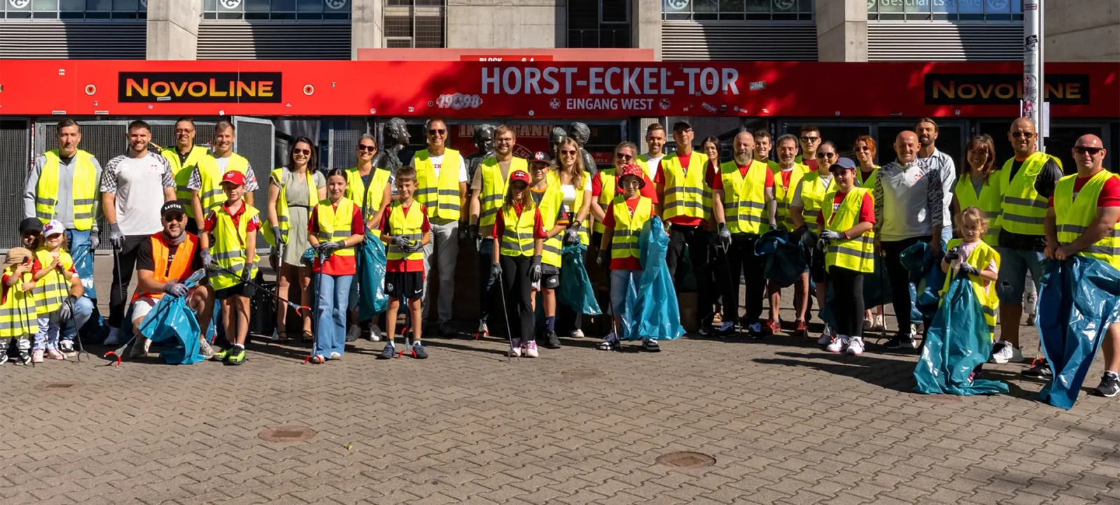 Eine Gruppe von Menschen, darunter Erwachsene und Kinder, steht vor dem Horst-Eckel-Tor eines Stadions und nimmt an einem Sauberkeitsprojekt teil. Sie tragen gelbe Sicherheitswesten und halten blaue Müllsäcke in den Händen. Das Bild zeigt eine gemeinsame Anstrengung zur Müllbeseitigung im Rahmen des World Cleanup Day.