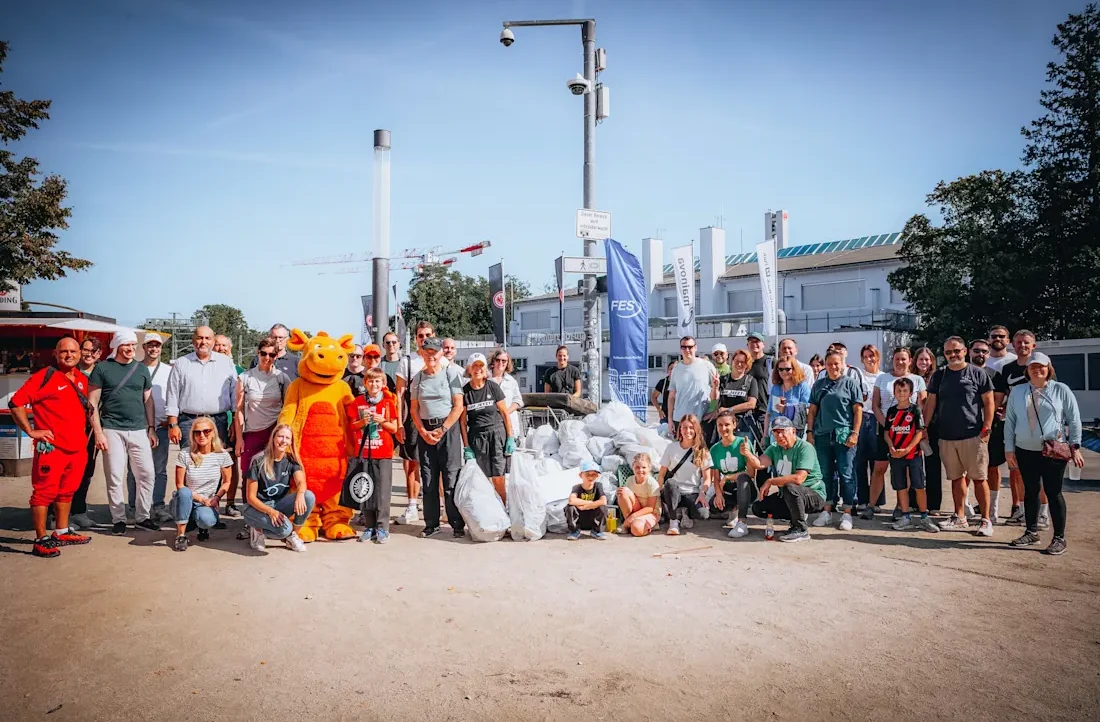 Eine Gruppe von Menschen, darunter auch Kinder, steht vor einem Stadion und hält Müllsäcke in den Händen. Sie haben an einem Clean-up-Day teilgenommen und sind stolz auf ihre Arbeit. Ein orangefarbenes Maskottchen steht in der Mitte der Gruppe. Im Hintergrund sind Bauten und Bäume zu sehen.