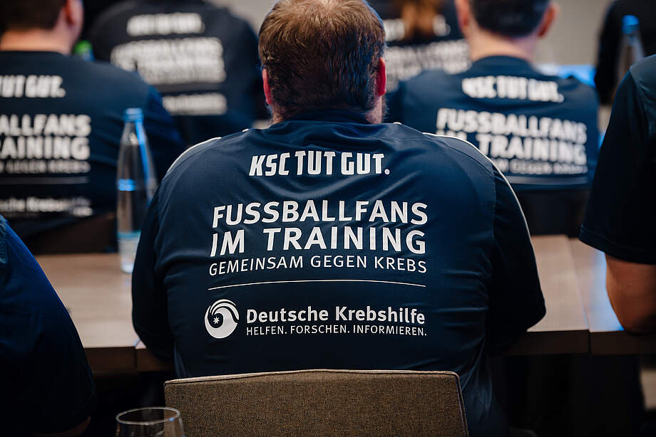 Mehrere Personen sitzen in einem Raum und tragen dunkle T-Shirts mit der Aufschrift "KSC TUT GUT. FUSSBALLFANS IM TRAINING GEMEINSAM GEGEN KREBS" und dem Logo der Deutschen Krebshilfe.