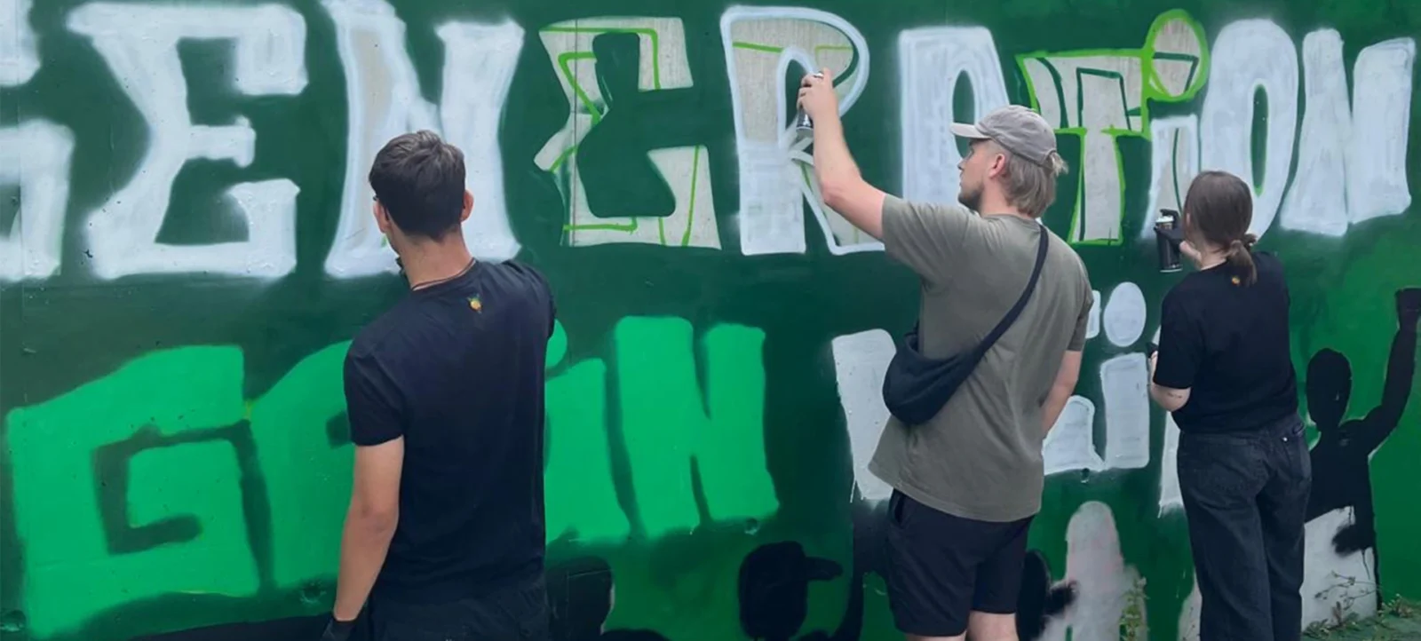 Drei Personen arbeiten gemeinsam an einem Graffiti-Kunstwerk. Eine Person malt mit einer Sprühdose, während die anderen beiden zuschauen und möglicherweise Anweisungen geben. Das Graffiti zeigt große, fett gedruckte Buchstaben in Weiß und Grün auf einem grünen Hintergrund. Die Szene findet im Freien statt, möglicherweise an einer Wand oder einem Zaun.