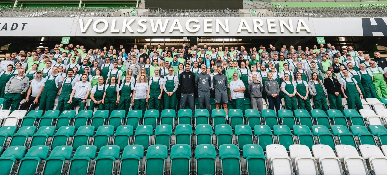 Eine große Gruppe von Menschen, darunter Spieler und Mitarbeiter des VFL Wolfsburg, steht in der Volkswagen Arena. Sie tragen einheitliche Kleidung, bestehend aus grauen T-Shirts und grünen Latzhosen. Die Gruppe ist vor grünen und weißen Sitzplätzen im Stadion positioniert. Im Hintergrund ist ein großes Schild mit der Aufschrift "Volkswagen Arena" zu sehen.