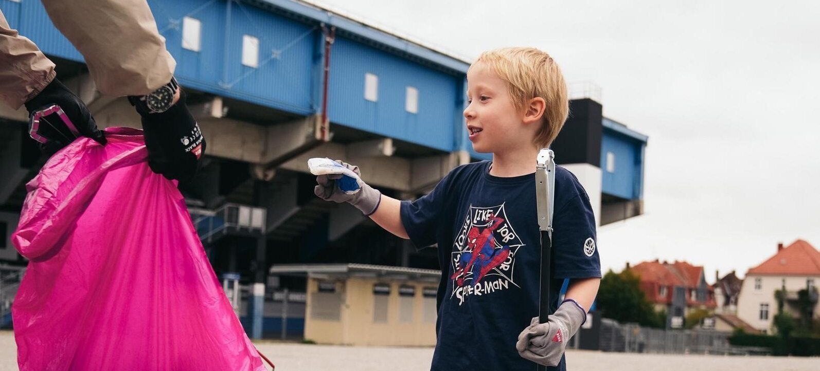 Ein junger Junge in einem Super-Man T-Shirt hält einem Erwachsenen einen Müllsammler hin, in den der Erwachsene gerade etwas Müll entsorgt. Der Junge trägt Handschuhe und hält einen Stock in der anderen Hand. Im Hintergrund ist ein blaues Stadiongebäude zu sehen.