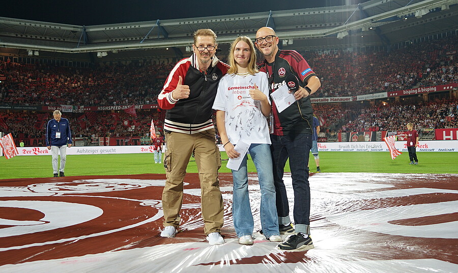Drei Personen stehen auf einem Fußballfeld und lächeln in die Kamera. Sie halten ein Schild mit der Aufschrift "Gemeinschaft fördert Leben". Im Hintergrund sind zahlreiche Zuschauer auf den Tribünen zu sehen. Die Personen tragen T-Shirts und Hoodies mit dem Logo des Vereins Hannover 96.