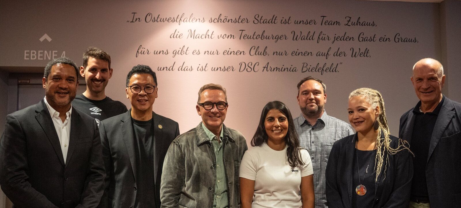 Gruppenfoto von Personen vor einer Wand mit einem Zitat über den DSC Arminia Bielefeld. Die Gruppe besteht aus sieben Männern und einer Frau, die lächeln und für das Foto posieren. Die Wand im Hintergrund trägt die Aufschrift "EBENE 4" und ein Zitat über die Schönheit der Stadt und die Verbundenheit mit dem DSC Arminia Bielefeld.