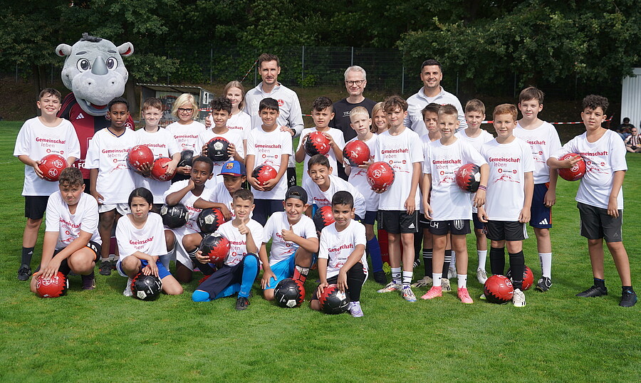 Eine Gruppe von Kindern und Erwachsenen steht auf einem Fußballfeld und hält rote und schwarze Fußbälle. Sie tragen weiße T-Shirts mit dem Aufdruck "Gemeinschaft" und "1. FC Köln". Im Hintergrund ist ein Maskottchen in Form eines Grauen Rhinozeros zu sehen. Die Szene wirkt fröhlich und Gemeinschaftsstiftend.