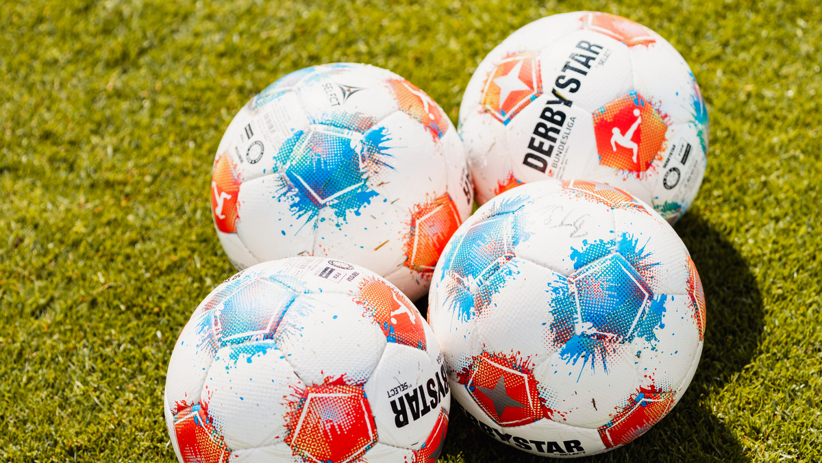 Vier Fußballbälle mit dem Design "Derbystar" liegen auf grünem Rasen. Die Bälle haben ein weißes Grunddesign mit roten und blauen Farbspritzern und einem Sternenlogo.