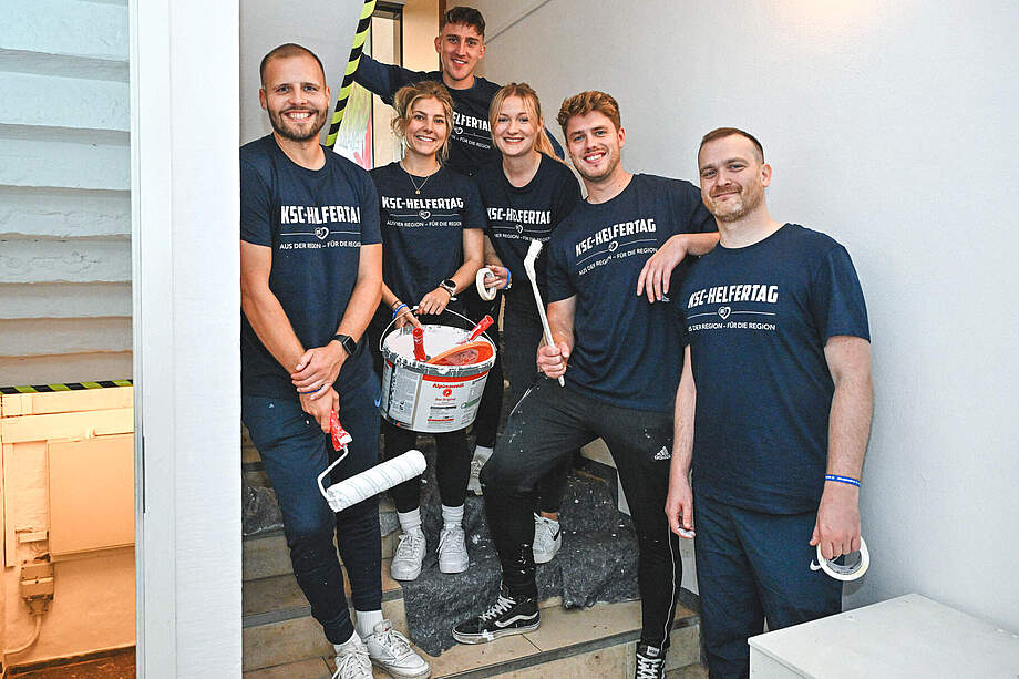 Eine Gruppe von sechs Personen steht auf einer Treppe und lächelt in die Kamera. Sie tragen alle schwarze T-Shirts mit der Aufschrift "KSC-Helfertag" und halten Malutensilien wie Farbrollen und einen Farbeimer. Die Szene wirkt fröhlich und engagiert, da sie offenbar an einem gemeinnützigen Projekt oder einer Renovierungsaktion teilnehmen.