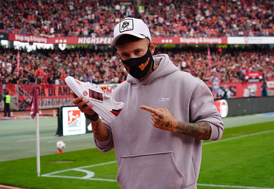 Ein Mann steht auf einem Fußballfeld und präsentiert ein Paar weiße Turnschuhe mit einem roten Logo. Er trägt eine graue Kapuzenjacke, eine weiße Baseballkappe und eine schwarze Maske mit der Aufschrift "Be Unique". Im Hintergrund sind viele Zuschauer auf den Tribünen zu sehen.