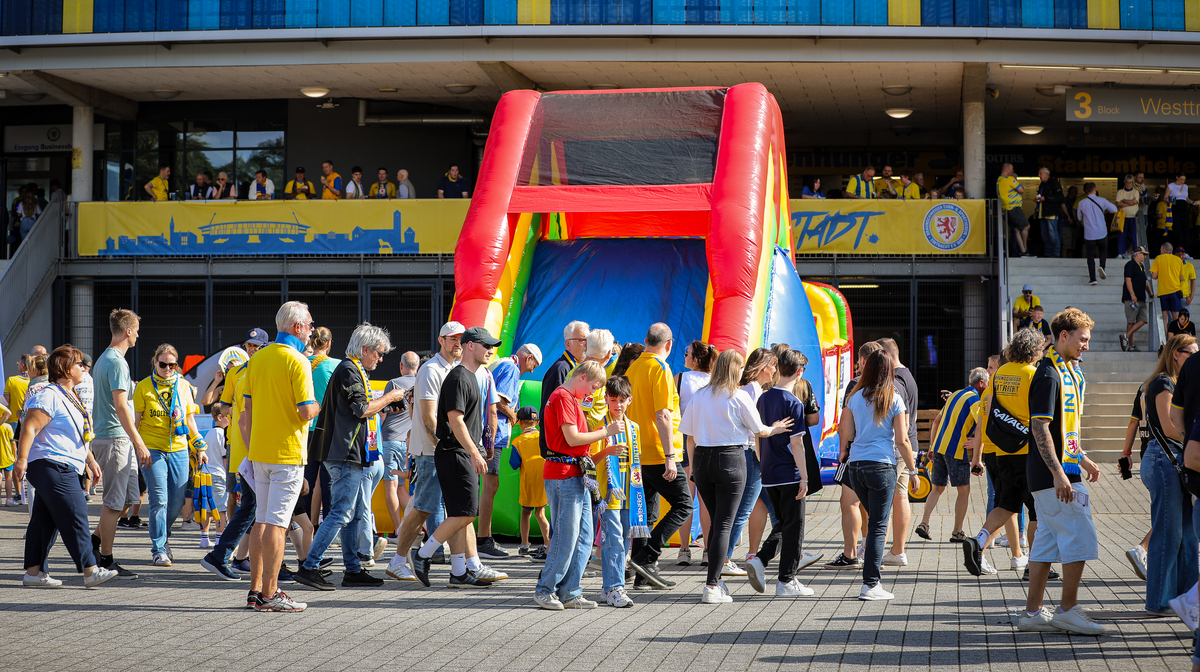 Eine Gruppe von Menschen, darunter Familien mit Kindern, versammelt sich um ein buntes, aufblasbares Spielgerät in Form eines großen Buchstabens 'H' vor einem Stadion. Die Menschen tragen verschiedene Kleidung, viele in gelb-schwarzen Farben, was auf eine Verbindung zu einem Fußballverein hindeutet. Im Hintergrund sind weitere Zuschauer auf den Stufen des Stadioneingangs zu sehen, und eine gelb-schwarze Banner mit einem Stadtsilhouetten-Motiv und einem Vereinslogo ist sichtbar.