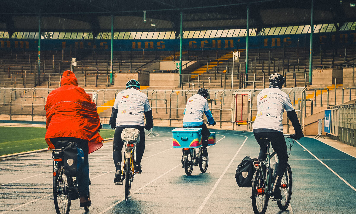 Vier Personen fahren mit Fahrrädern auf einer Laufbahn in einem Stadion. Eine Person trägt eine rote Jacke und die anderen drei tragen weiße T-Shirts mit verschiedenen Logos. Die Fahrräder haben Taschen und Gepäck. Im Hintergrund sind leere Tribünen zu sehen.